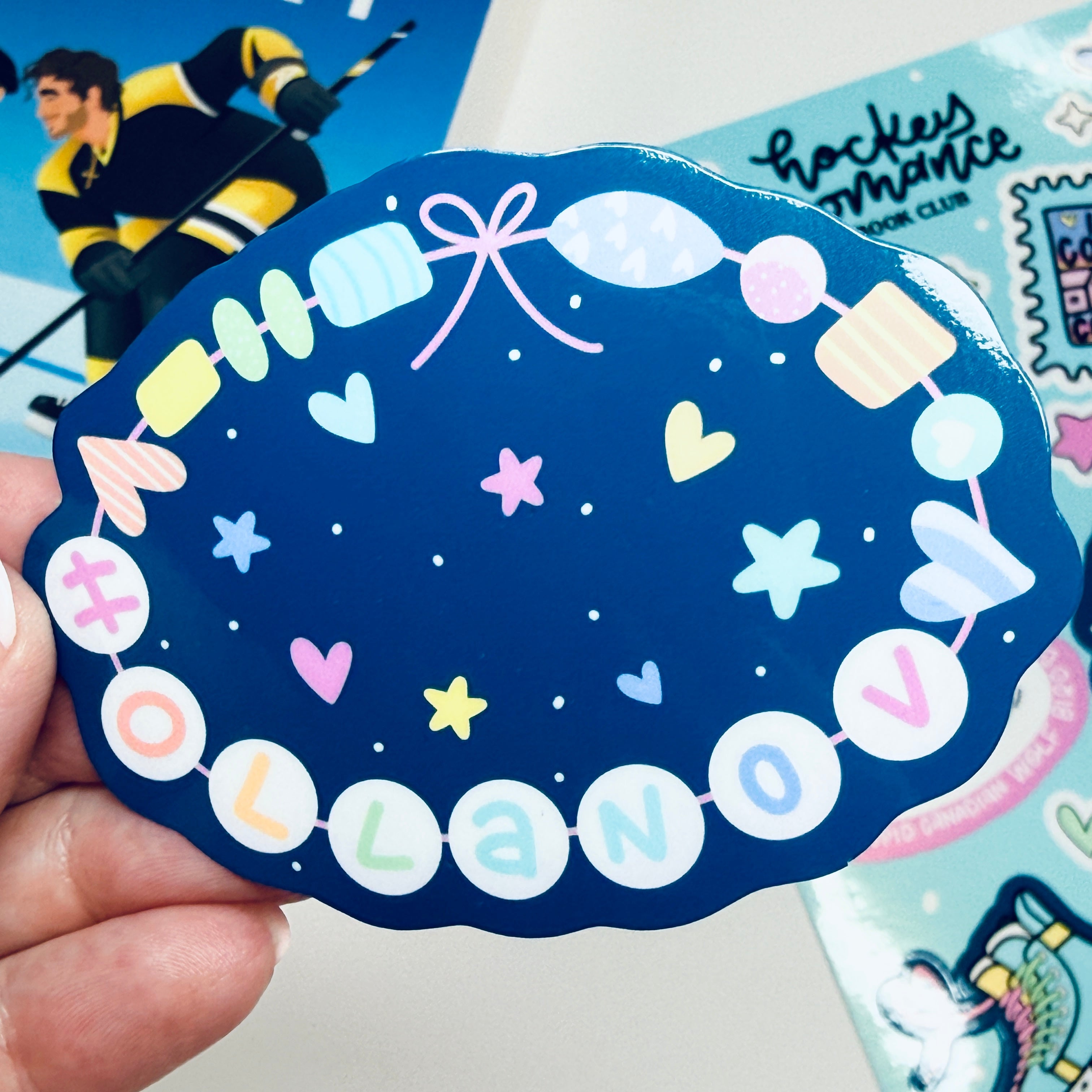 Decorative sticker with colorful letters and designs held by a hand describing Hollanov on a friendship bracelet